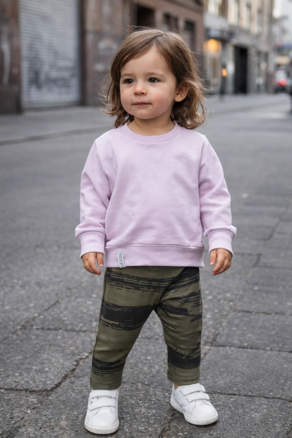 Tragebild einen mädchens ca 2 jahre mit dunklen haaren die eine oliv farbene leggings mit schwarzen pinselstrichen und einen sweater in hellem lila trägt dazu weiße sneaker im Hintergrund eine urbane szene
